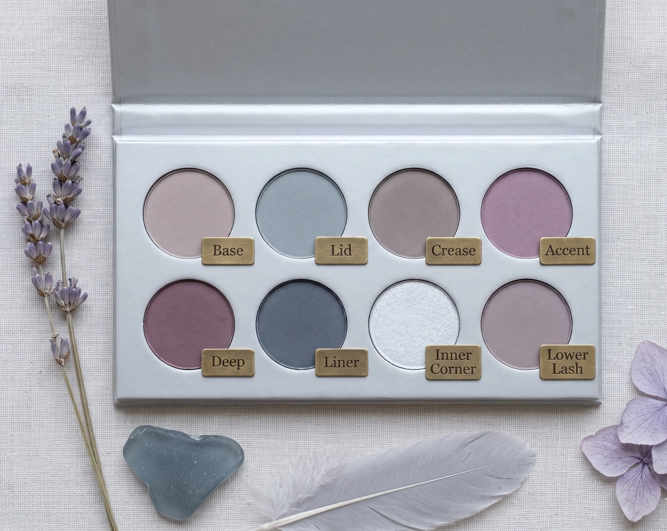 Soft Summer eyeshadow palette arranged in a silver compact tray showing eight cool muted shades — greyed taupe, dusty mauve, slate grey, cool pearl, dusty plum — with corresponding natural objects: dried lavender, sea glass, and pale hydrangea