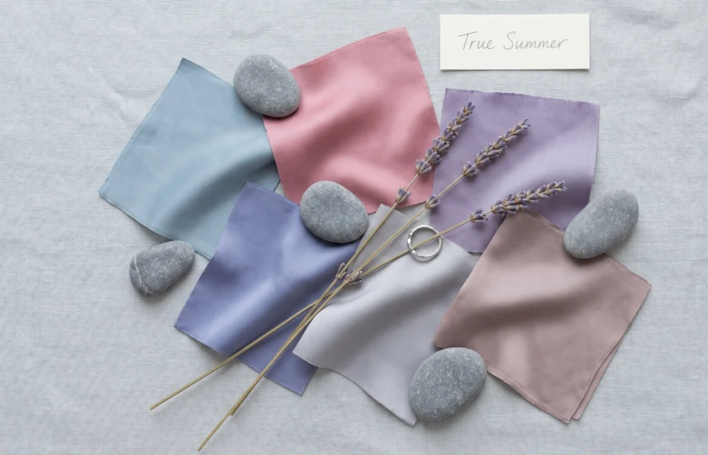 True Summer colour palette flat-lay — silk swatches in misty blue, cool rose pink, muted lavender and dove grey with river stones and dried lavender, representing the true summer celebrity colour season