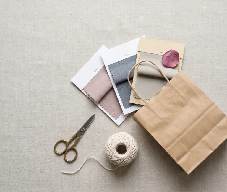 A shopping bag with dusty rose, slate blue-grey, and cool greige fabric swatches spilling out — a visual reference for shopping soft summer clothing brands and stores.