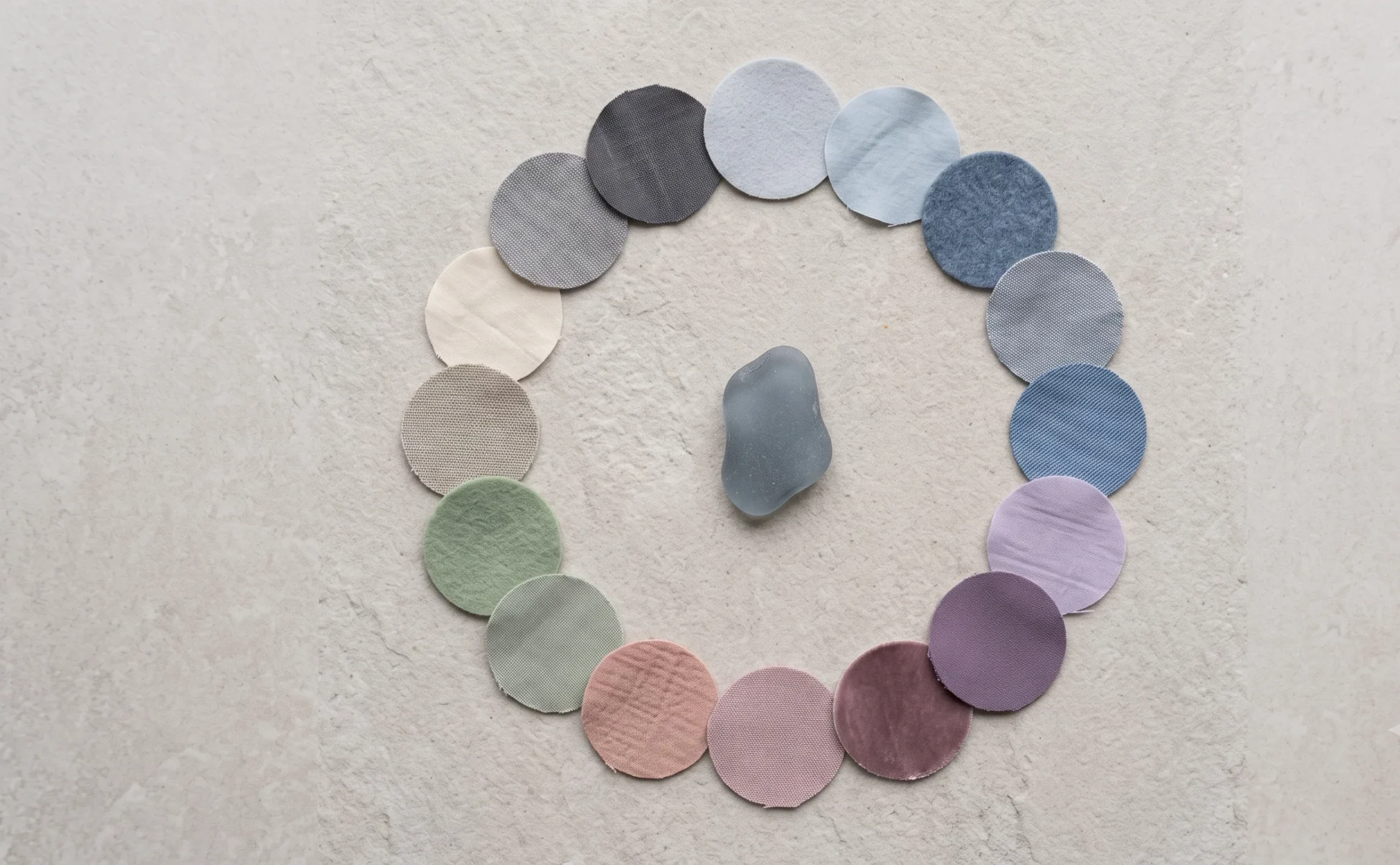 Sixteen circular fabric swatches arranged in a radial wheel formation representing the Soft Summer colour wheel, with a piece of sea glass at the centre, showing the full palette's relationship from cool neutrals through blues, purples, pinks, and greens