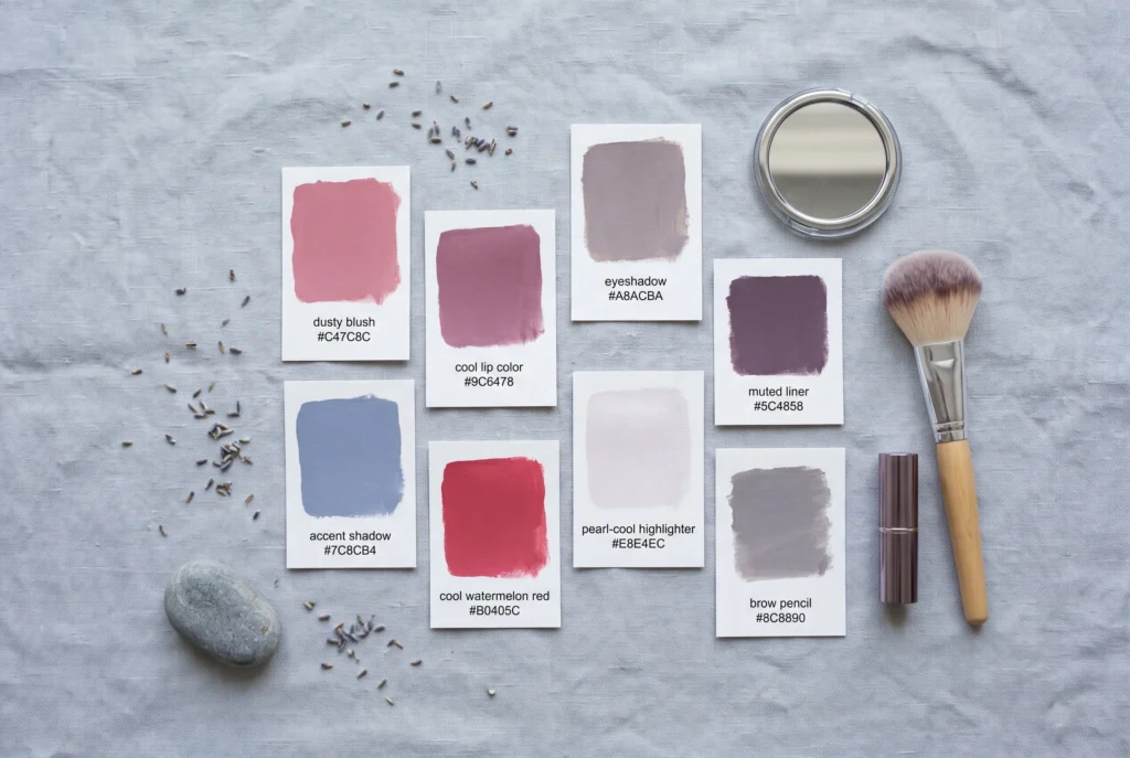 True Summer makeup colour palette flat-lay — cosmetic swatch cards in dusty rose, cool mauve, taupe-grey and slate blue with a compact mirror and lipstick bullet, showing the cool summer makeup colour range